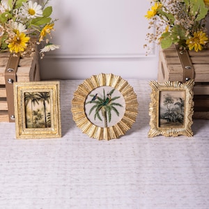 May include: Three gold-toned picture frames with ornate designs. The frames are arranged in a row on a white surface. The center frame features a sunburst design and a picture of a palm tree with two birds. The frames on either side feature a more traditional design and pictures of palm trees.