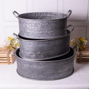 May include: Three galvanised metal oval tubs with handles, stacked together. The tubs are of varying sizes and have a hammered texture. The handles are made from the same metal. The largest tub is at the base of the stack, with the others nested inside.