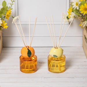 May include: Two glass reed diffusers with orange and lemon ceramic toppers. The orange diffuser has a textured orange base with a green leaf, while the lemon diffuser is yellow with a small green leaf. Both contain amber liquid and wooden reeds.