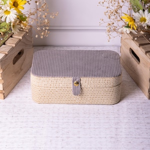 Op de afbeelding: Een opbergdoos van wilgenvlechtwerk met een grijze fluwelen voering en een gouden knoopsluiting.