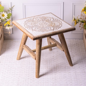 May include: A small, square wooden stool with a white top featuring a beige, ornate mandala design. The stool has four angled legs and a supportive crossbar. The wood has a natural, light brown finish. The stool is likely used for decorative purposes.