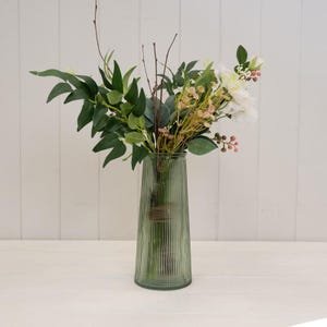 May include: A tall, ribbed, sage green glass vase containing an arrangement of artificial greenery, thin brown twigs, small pink berry clusters, and white flowers. The vase has a tapered cylindrical shape with vertical ridges along the exterior.