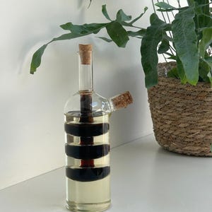 Könnte beinhalten: Eine klare Öl- und Essigflasche aus Glas mit einem Korken. Die Flasche hat eine zylindrische Form mit drei schwarzen Bändern in der Mitte. Die Flasche ist mit einer klaren Flüssigkeit gefüllt. Ein zweiter Ausguss mit einem Korken ist sichtbar.