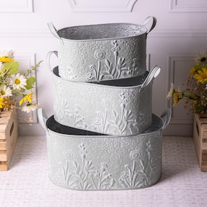 May include: Three oval, light grey metal storage bins stacked together. Each bin has embossed floral patterns and curved handles. These decorative bins are suitable for home storage and decor, and are ideal for any room.