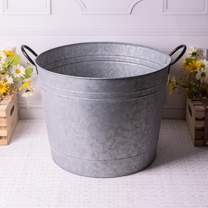 May include: A galvanised metal bucket with two black handles. The bucket is oval-shaped and has three horizontal bands around the body. The bucket is empty and sits on a white surface. Decorative flowers are in wooden crates on either side.