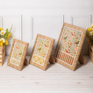 May include: Three rectangular picture frames with light brown wood-grain finish, each containing a print featuring stylised birds, leaves, and floral patterns in shades of green, orange, and yellow, displayed in ascending size order.