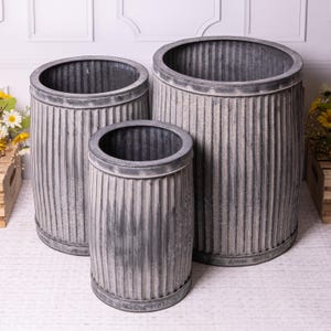 May include: A set of three galvanised metal planters in different sizes. They have a ribbed texture and a weathered grey finish. Each planter has a rounded rim and a cylindrical shape, suitable for indoor or outdoor use.