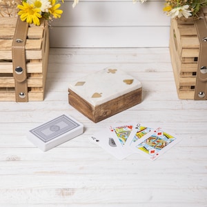 Peut inclure: Une boîte à cartes en bois avec un dessus en marbre blanc incrusté de symboles de jeu dorés. Un jeu de cartes et plusieurs cartes à jouer sont affichés sur une surface en bois blanc. Deux caisses décoratives en bois sont en arrière-plan.