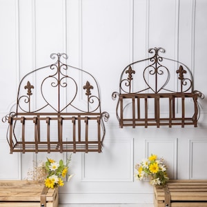 May include: Two decorative, brown metal wall planters featuring fleur-de-lis and heart motifs. Each planter has a rectangular basket for plants. The planters are mounted on a white wall, with flowers in wooden crates below.