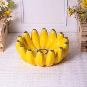 Può includere: Una ciotola in ceramica giallo brillante a forma di grappolo di banane. La ciotola ha una finitura lucida e punte marroni su ogni banana. Un oggetto decorativo ideale per la cucina o la sala da pranzo.