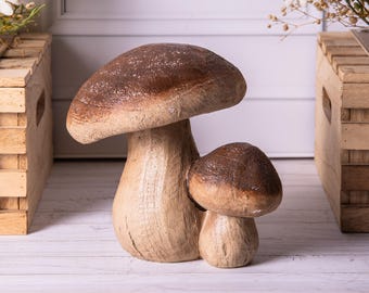 Könnte beinhalten: Zwei dekorative Pilzskulpturen. Der größere Pilz hat einen hellbraunen Stiel und einen dunkelbraunen Hut. Der kleinere Pilz hat ein ähnliches Farbschema. Die Skulpturen bestehen aus einem strukturierten Material und stehen auf einer weißen Oberfläche.