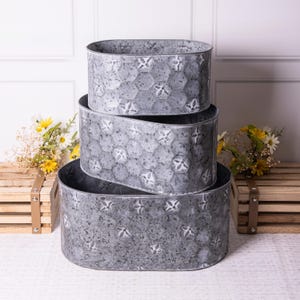 May include: A set of three oval galvanized metal planters. The planters are grey with a textured surface and a bee pattern. The planters are nested, with the largest at the bottom and the smallest at the top.