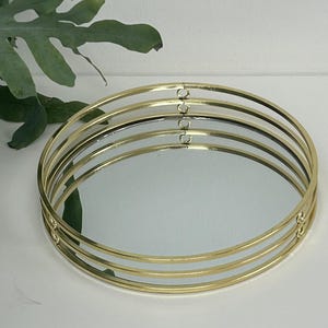 Könnte beinhalten: Ein runder Dekoteller mit verspiegelter Oberfläche und einem goldfarbenen Metallrahmen. Der Rahmen besteht aus mehreren konzentrischen Ringen, die einen geschichteten Effekt erzeugen. Der Teller dient zur Präsentation von Gegenständen und hat eine schlichte, elegante Ästhetik.