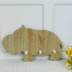 Könnte beinhalten: Schlüsselhalter in Form eines Holz-Nilpferds mit drei weißen Haken. Der Schlüsselhalter besteht aus hellfarbenem Holz mit sichtbarer Maserung und einem lächelnden Gesicht.