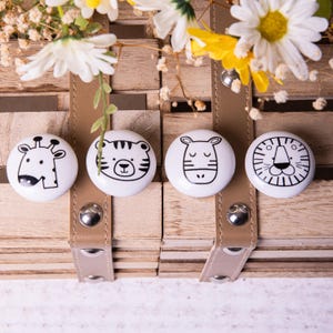 May include: Four white ceramic knobs with black animal illustrations. The knobs feature a giraffe, a tiger, a zebra, and a lion. The knobs are arranged in a row on a wooden surface.