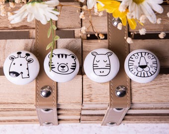 May include: Four white ceramic knobs with black animal illustrations. The knobs feature a giraffe, a tiger, a zebra, and a lion. The knobs are arranged in a row on a wooden surface.