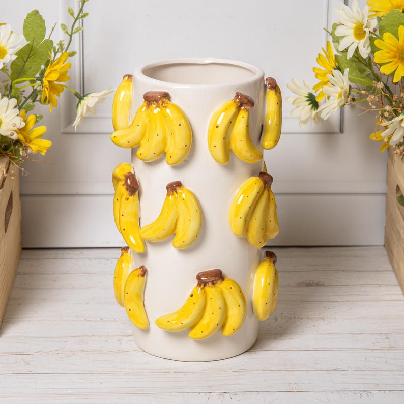 Peut inclure: Un vase en c&eacute;ramique blanc de forme cylindrique, orn&eacute; de grappes de bananes jaunes en relief et de tiges marron. Le vase est un objet d&eacute;coratif, parfait pour contenir des fleurs ou comme pi&egrave;ce d'accent.