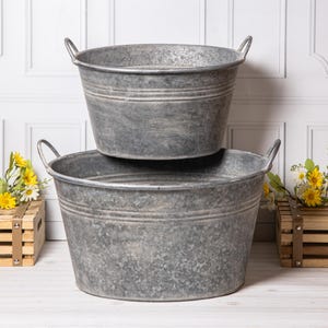 May include: Two round, galvanised metal tubs with a weathered, mottled grey finish. Each tub features two riveted metal handles on opposite sides and horizontal ribbed detailing around the upper circumference, stacked one on top of the other.