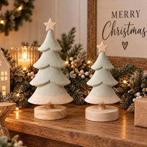 May include: Two wooden Christmas tree decorations with a pale green finish on the branches and natural wood bases. Each tree features a carved star topper and a tiered design, standing on a circular wooden pedestal for a stable display.