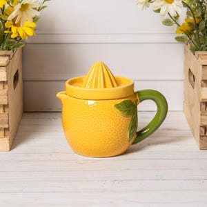 Może przedstawiać: Żółta ceramiczna wyciskarka do cytrusów w kształcie cytryny o teksturowanej powierzchni, ze spiczastą pokrywką i zakrzywionym zielonym uchwytem. Korpus wyciskarki posiada wyrzeźbiony detal zielonego liścia z boku, stanowiąc funkcjonalne i dekoracyjne narzędzie kuchenne.