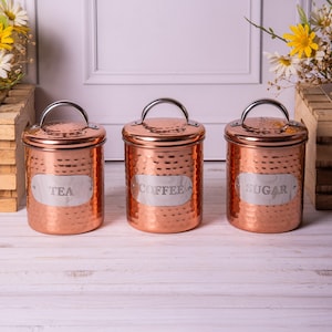 May include: Three copper-coloured canisters with silver handles and lids. Each canister has a hammered texture and a silver label with the words "TEA", "COFFEE", and "SUGAR". The canisters are arranged on a light wood surface.