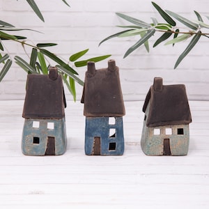 May include: Three ceramic house ornaments with brown roofs and blue-green walls. Each house has cut-out windows and a door. The ornaments are arranged on a white wooden surface, with green foliage in the background.