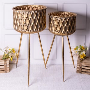 May include: Two gold metal plant stands with a diamond pattern. The stands have three legs and are made of metal.