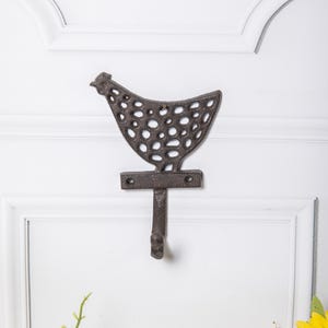 Puede incluir: Un gancho de pared decorativo de hierro fundido con forma de gallina. El gancho es de color marrón oscuro con un acabado envejecido y un cuerpo perforado. Está montado en una puerta blanca.