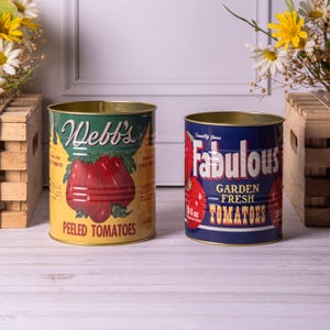 Może przedstawiać: Dwa metalowe puszki w stylu vintage. Jedna jest żółta z napisem "Webb's Peeled Tomatoes" i ilustracją pomidora. Druga jest niebieska z napisem "Fabulous Garden Fresh Tomatoes" i grafiką pomidorów. Obie puszki są otwarte u góry.