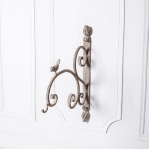 May include: A decorative, weathered metal wall-mounted bracket with a bird and scroll design. The bracket features a small bird perched on a curved arm, with decorative scrollwork and leaf accents. The overall color is a muted, antique-style gray.