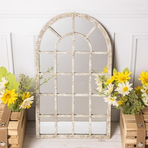 Peut inclure: Un miroir mural en forme de fenêtre arquée avec un cadre blanc cassé vieilli et un motif en grille. Le miroir est flanqué de deux caisses en bois contenant chacune un bouquet de marguerites jaunes et blanches avec du feuillage vert.