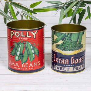 Peut inclure: Deux boîtes de conserve cylindriques en métal avec des étiquettes de style vintage. La boîte rouge à gauche présente des illustrations de haricots de Lima et le texte POLLY BRAND LIMA BEANS. La boîte bleue à droite montre des petits pois.