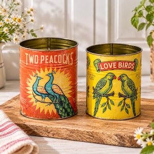 Pode incluir: Duas latas metálicas cilíndricas com rótulos de estilo vintage. A lata laranja maior apresenta dois pavões azuis e o texto "TWO PEACOCKS". A lata amarela menor mostra dois papagaios verdes num galho com o texto "LOVE BIRDS".