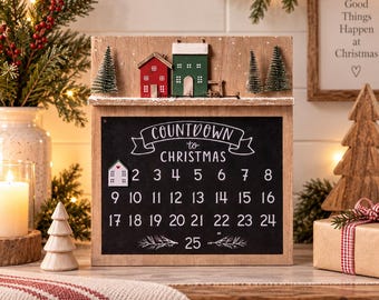 Puede incluir: Un calendario de cuenta regresiva navideña de madera con una superficie de pizarra negra, números del 1 al 25 impresos en blanco y el texto Countdown to Christmas. El estante superior muestra una casa roja, una verde y tres pinos en miniatura.
