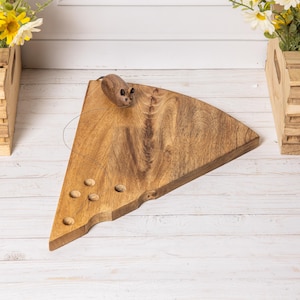 Könnte beinhalten: Ein dreieckiges Servierbrett aus Holz in Form eines Käsestücks mit natürlicher Holzmaserung und mehreren kleinen kreisförmigen Vertiefungen. Eine kleine, geschnitzte Holzmausfigur ist nahe der oberen Kante des Brettes positioniert.