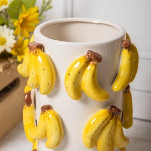 Peut inclure: Un vase en c&eacute;ramique cr&egrave;me orn&eacute; de grappes de bananes jaunes. Les bananes ont des tiges marron et de petites taches noires. Le vase cylindrique a une finition lisse et brillante. Le design est fantaisiste et d&eacute;coratif.