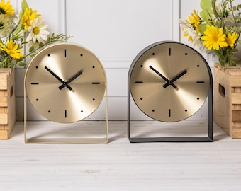 Op de afbeelding: Twee ronde tafelklokken met metallic gouden wijzerplaten en zwarte wijzers. De ene klok heeft een goudkleurig frame, terwijl de andere een matzwart frame heeft. Beide klokken tonen eenvoudige zwarte uurmarkeringen en staan rechtop.