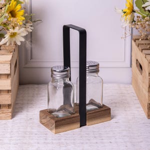 May include: A clear glass salt and pepper shaker set with a stainless steel top and a black metal handle. The set sits on a wooden base. The background includes wooden crates and floral arrangements.