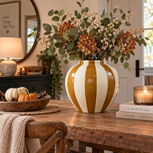 May include: A round ceramic vase featuring vertical stripes in mustard yellow and off-white. The vase has a glossy finish and is filled with a large arrangement of dried eucalyptus branches, small white flowers, and rust-coloured hydrangea blooms.