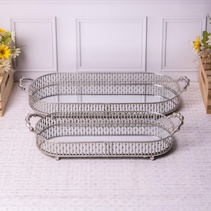 May include: Two silver oval trays with mirrored bases. Each tray features decorative handles and a detailed border design. The trays are of different sizes and are placed on a white surface.