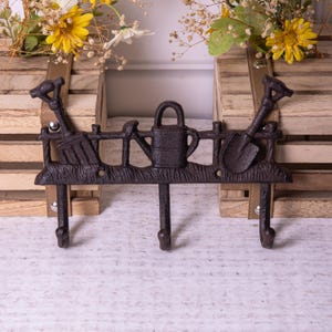 May include: A cast iron wall mounted coat rack with three hooks. The rack is decorated with a fence, a watering can, a shovel, and a rake. The rack is brown in colour.