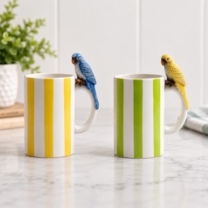 May include: Two ceramic mugs featuring vertical stripes and a sculpted bird handle. The left mug has yellow stripes and a blue parakeet handle, while the right mug has green stripes and a yellow parakeet handle. Both mugs have white interiors.
