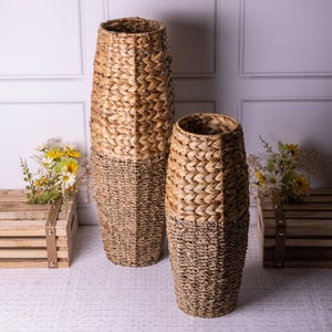 May include: Two woven wicker vases, one tall and one shorter, both in a natural brown colour. The vases are made from a woven material and have a rustic look.