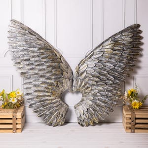 May include: Large, decorative metal angel wings with a heart-shaped cutout in the centre. The wings are silver and gold, with a detailed feather design. The wings are displayed against a white wall.