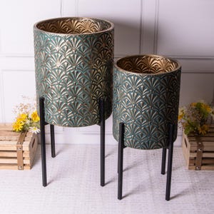May include: Two decorative plant pots. The pots are cylindrical with a teal and gold art deco pattern. They are supported by black metal stands. The pots are different sizes and are empty.