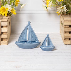 Op de afbeelding: Twee blauwe keramische zeilbootfiguren. De zeilboten hebben een uitgesneden zeilontwerp en staan op een wit houten oppervlak. Gele en witte bloemen in houten kisten staan op de achtergrond.