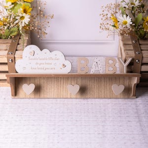 Op de afbeelding: Houten plank met een wit wolkenbord waarop staat "Twinkle twinkle little star, do you know how loved you are." De plank heeft ook het woord "BABY" gespeld in houten letters met witte stippen. Er zijn drie hartvormige haken op de plank.