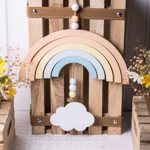 May include: A wooden rainbow mobile with pastel colors hanging from a wooden crate. The rainbow is made of five arches in shades of peach, yellow, light blue, and white. A white cloud hangs from the bottom of the rainbow with a string of pastel beads.