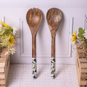 Op de afbeelding: Een houten saladeservies bestaande uit een lepel en een vork. De handvatten zijn donkerbruin en de uiteinden zijn versierd met een wit patroon van groene bladeren en zwarte olijven. De set wordt gepresenteerd tegen een witte achtergrond.