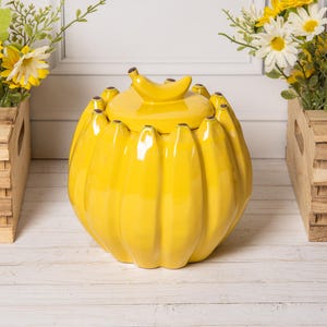 May include: A bright yellow ceramic jar shaped like a cluster of bananas. The jar features a rounded body with vertical ridges mimicking individual banana peels and a matching lid topped with a small, curved banana-shaped handle for easy lifting.